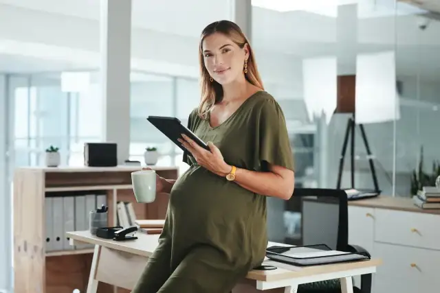 Uśmiechnięta ciężarna kobieta z tabletem i kubkiem w biurze. Dowiedziałam się o ciąży po okresie wypowiedzenia, ale teraz planuję przyszłość.