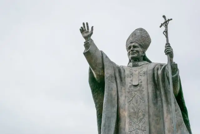 Pomnik Jana Pawła II z podniesioną ręką i krzyżem. Wierni odmawiają modlitwę do Jana Pawła II o uzdrowienie.