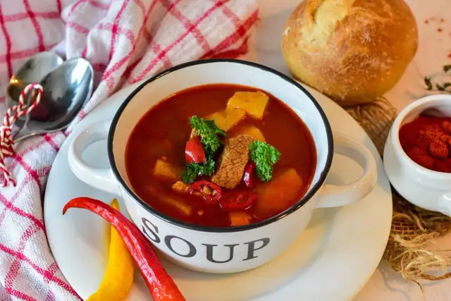 Miska gulaszowej zupy z mięsem, ziemniakami i papryczkami chili, obok chleb i przyprawy.