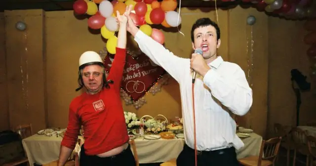 Dwóch mężczyzn, jeden w białym kasku i czerwonej koszulce, drugi z mikrofonem, świętuje. W tle balony i dekoracje weselne.