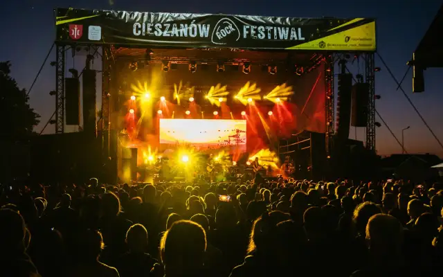 Tłumy bawią się na Cieszanów Rock Festiwal. Jasne światła sceny oświetlają zespół grający na tle zachodzącego słońca.