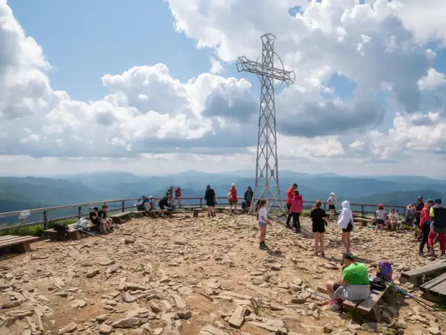 Ludzie odpoczywają na szczycie, podziwiając widoki na Bieszczady. Metalowy krzyż góruje nad kamienistym terenem, a w oddali widać łagodne pasma górskie.