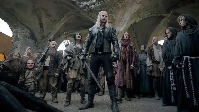 Geralt z Rivii stoi pośród tłumu, gdy ścieżki przeznaczenia w Wiedźminie splatają się. Widać krasnoludy, ludzi i zakapturzonych mnichów.