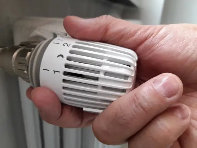 Dłoń reguluje termostat grzejnika, pokazując jak zakręcić zawór grzejnika, by oszczędzać ciepło.
