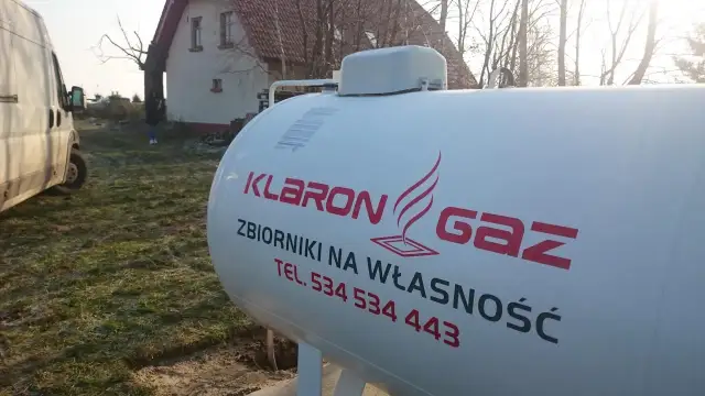Biały zbiornik na gaz z logo firmy Klaron Gaz, gotowy do wymiany butli LPG. W tle dom i biały van.