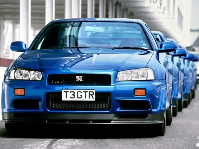 Błękitny Nissan Skyline GT-R R34 z tablicą "T3 GTR" ustawiony w rzędzie z innymi.