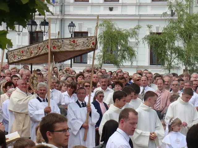 Procesja religijna z monstrancją, tłum ludzi w białych szatach, budynek w tle.