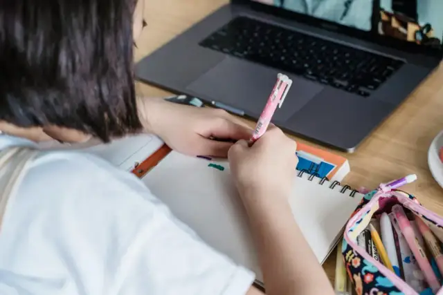 Dziewczynka pisze w zeszycie, obok leży laptop i piórnik z kredkami.