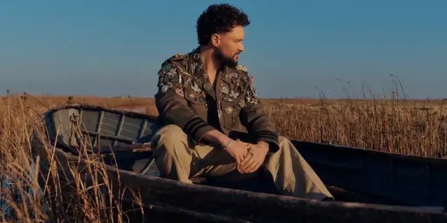 Calum Scott w łodzi, w kurtce ozdobionej broszkami, jakby śpiewał "You Are The Reason".