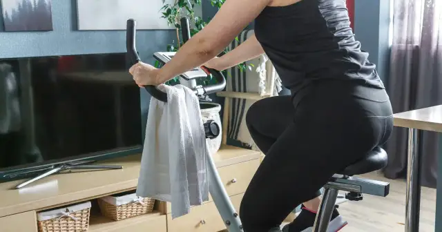 Kobieta ćwiczy na rowerku stacjonarnym, osiągając efekty w domowym zaciszu. Obok ręcznik i telewizor.
