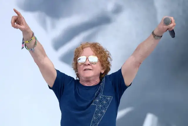 Wokalista Simply Red z uniesionymi rękami, trzymający mikrofon. Ma charakterystyczne rude włosy i okulary przeciwsłoneczne.