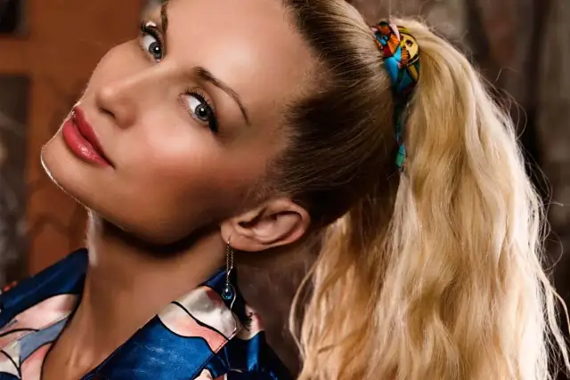Kobieta z blond włosami spiętymi w kitkę z przedziałkiem, ozdobioną kolorową chustką.