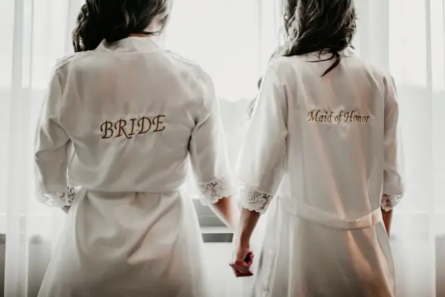 Dwie kobiety w szatach, jedna z napisem "BRIDE", druga "Maid of Honor". Idealny obrazek, by zainspirować się, jak poprosić świadka na ślub.