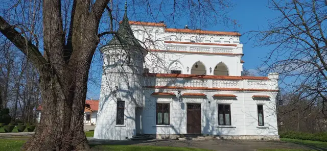 Biały pałac z wieżyczką i czerwonym dachem, otoczony drzewami. W oddali widać inne budynki. To piękny widok na **Radomyśl Wielki**.