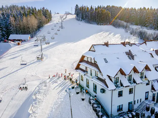 Hotel ze stokiem narciarskim dla dzieci. Kolejka krzesełkowa wwozi narciarzy na ośnieżony stok, a obok hotelu bawią się dzieci.