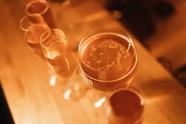 Kilka kieliszków z bursztynowym napojem, jeden z nich z widocznym napisem "kombucha".