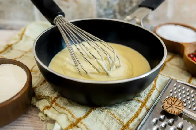 Eine cremige low carb bechamel wird in einer Pfanne mit einem Schneebesen verrührt. Daneben stehen Schüsseln mit Sahne, Salz und Pfefferkörnern.