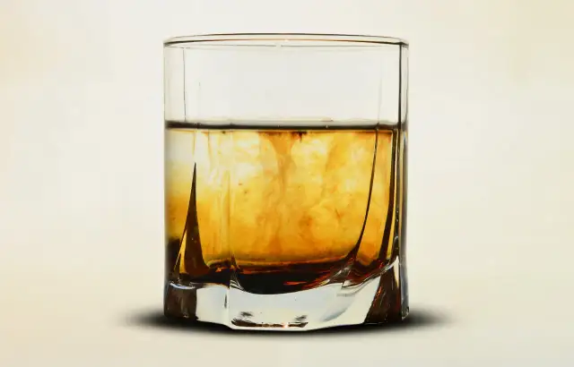 Szklanka z bursztynowym płynem, w którym rozpuszcza się ciemniejsza substancja, przypominająca proces, jak zrobic whiskey.