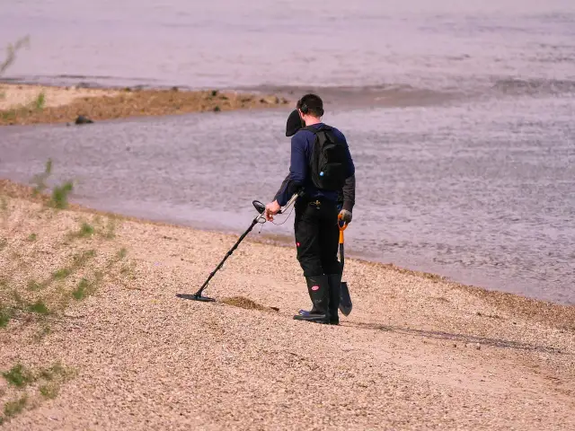 Mężczyzna z wykrywaczem metali i łopatką na plaży przy wodzie.