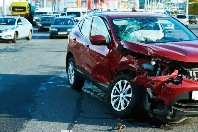 Wypadek drogowy. Czerwony samochód po kolizji, zniszczony przód i pęknięta szyba. Na drodze inne pojazdy, w tym radiowóz. Może to być sytuacja wymagająca **umowy na zastępstwo** kierowcy.