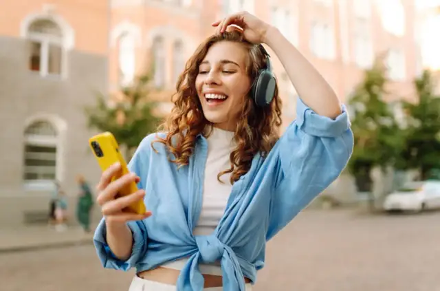 Kobieta w słuchawkach i niebieskiej koszuli trzyma żółty telefon, słuchając muzyki.