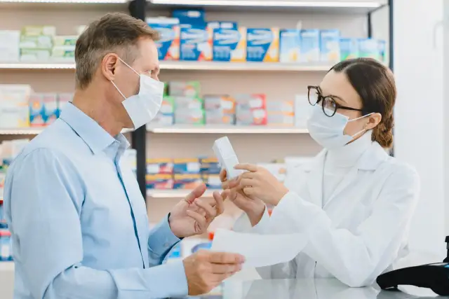 Farmaceutka w masce i okularach podaje pacjentowi w masce pudełko leku.