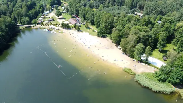 Słoneczny dzień nad jeziorem Rogoźnik. Ludzie kąpią się i opalają na piaszczystej plaży, otoczonej zielonym lasem.