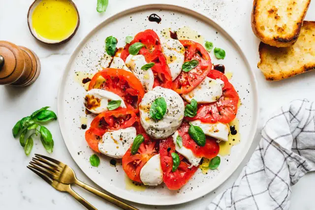 Caprese-Salat mit frischen Tomaten, veganem Mozzarella und Basilikum, beträufelt mit Olivenöl und Balsamico. Dazu geröstetes Brot.