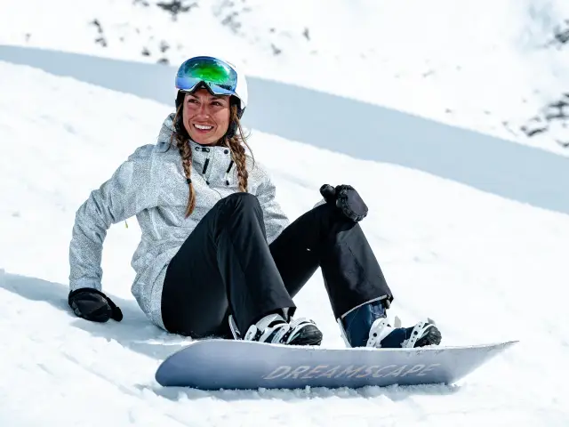 Uśmiechnięta snowboardzistka w kasku i goglach siedzi na śniegu obok deski. Jej pokrowiec na deskę snowboardową jest gotowy do drogi.