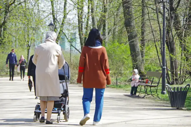 Kobieta z wózkiem spaceruje po parku, w tle inne osoby i dziecko na ławce.