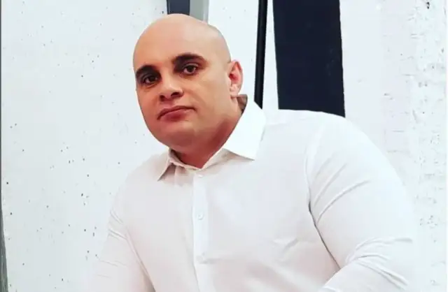 Dominik Abus, wiek nieznany, w białej koszuli, na tle białej ściany z czarnymi pasami.