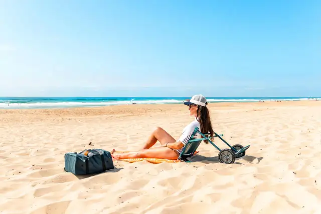 Kobieta w czapce i okularach przeciwsłonecznych siedzi na plaży na składanym krześle obok torby.