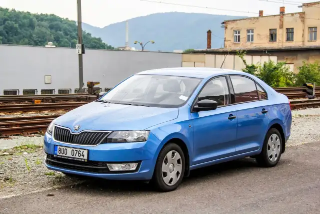 Niebieski sedan Skoda Rapid stoi przy torach kolejowych. W tle budynki i góry.