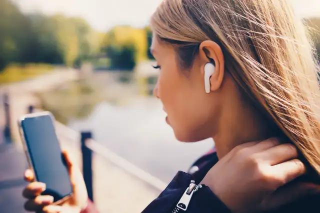 Kobieta z blond włosami w białych słuchawkach bezprzewodowych trzyma telefon.