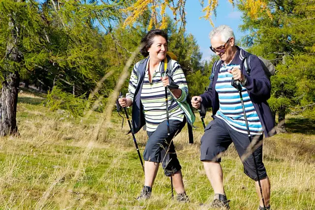 Starsza para wesoło wędruje po lesie z kijkami trekkingowymi.