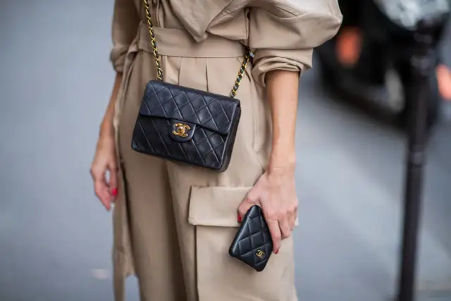 Elegancka kobieta w beżowym płaszczu prezentuje klasyczną, pikowaną torebkę Chanel vintage z złotym łańcuszkiem i logo.