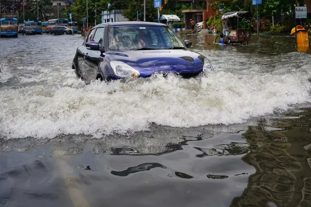 Niebieskie auto walczy z falą wody na zalanej ulicy. W tle widać inne auta po powodzi i autobusy.