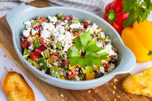 Bunter Paprikasalat mit Feta, garniert mit Petersilie, serviert in einer hellblauen Schüssel auf einem Holzbrett. Daneben liegen geröstete Brotscheiben.