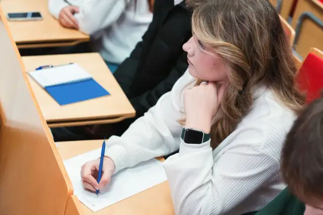 Studentka pisze notatki na wykładzie, opierając podbródek na dłoni.