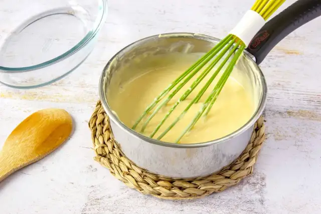 Un batidor verde mezcla una crema amarilla en una olla, mostrando cómo espesar un puré. Cerca hay una cuchara de madera y un recipiente de vidrio.