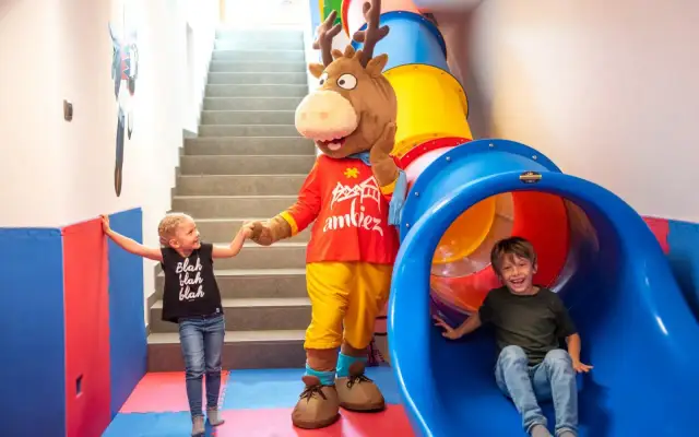 Mascot and two children in a colorful indoor playground with a slide and stairs.