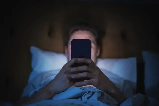 Mężczyzna leżący w łóżku i patrzący na ekran telefonu w ciemności.