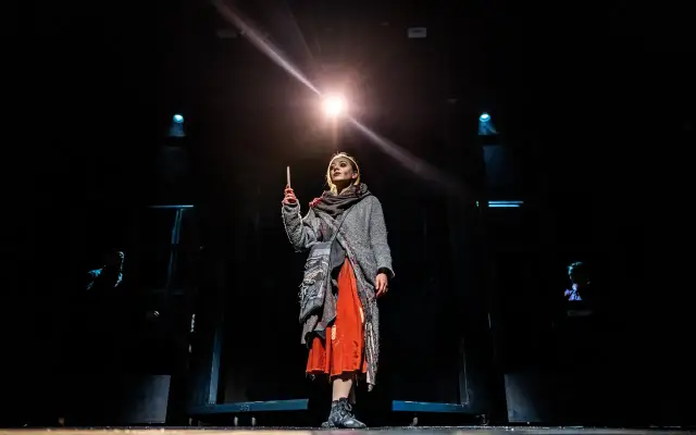 Dziewczynka z zapałkami na scenie, trzymająca płonącą zapałkę, w otoczeniu mroku i teatralnego światła.