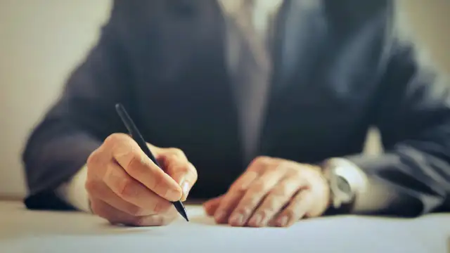 Dłoń w garniturze pisząca długopisem na białym papierze.