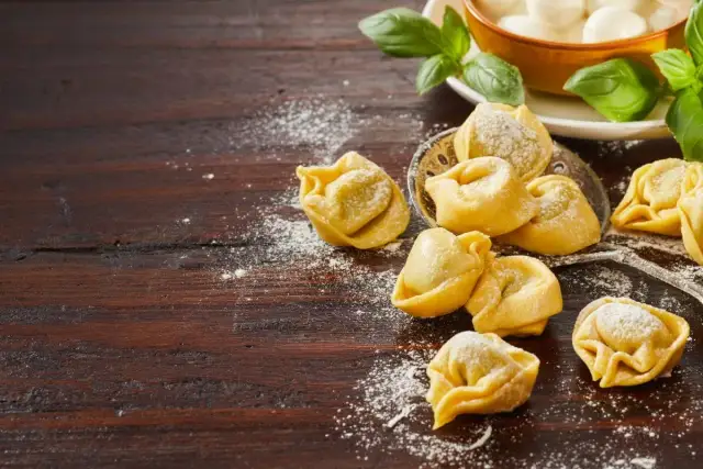Surowe tortellini posypane mąką na drewnianym blacie, obok liście bazylii i miseczka z mozzarellą.