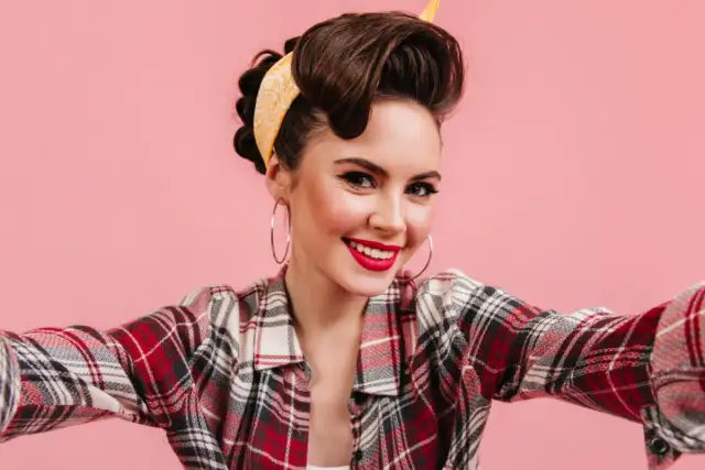 Uśmiechnięta kobieta w stylu pin-up, z burzą brązowych włosów spiętych w kok i żółtą bandaną, w kratę koszuli, robi sobie selfie.