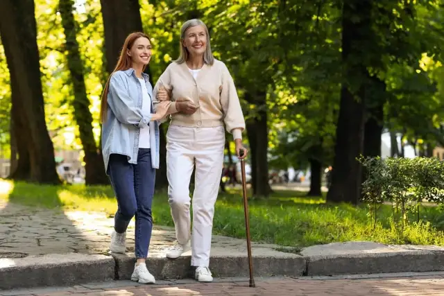 Młoda kobieta pomaga starszej pani, która wspiera się na czwórnodze, spacerując po parku.