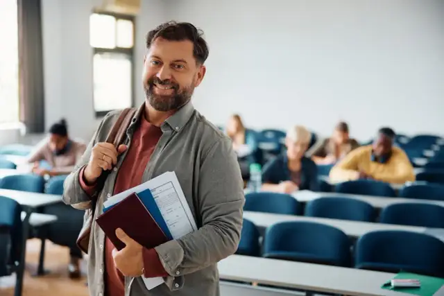 Uśmiechnięty mężczyzna z teczką w ręku, w tle sala wykładowa. Czy po technikum można iść do szkoły policealnej? Tak, to świetna ścieżka rozwoju!