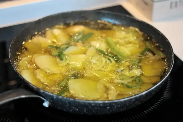 Patatas panaderas en sartén, dorándose en aceite caliente con cebolla y hierbas.