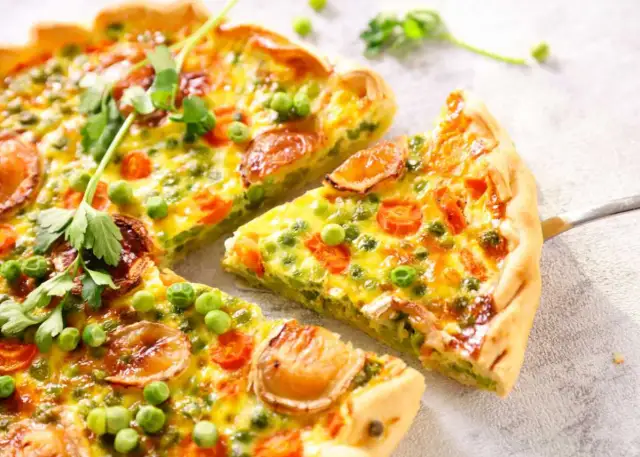 Un delicioso quiche de verduras, con guisantes, zanahoria y trozos de queso dorado, recién cortado y listo para servir.